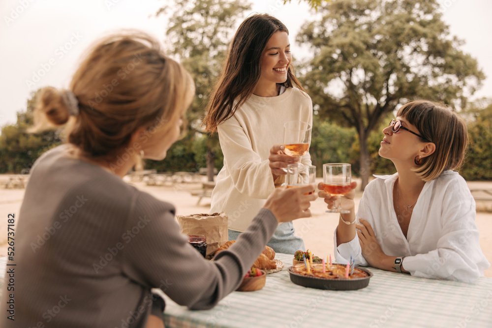 Cute young caucasian women at table celebrate first day of summer at picnic in park. Girlfriends hold glasses with booze, wear casual clothes. Concept enjoying weekend, vacation Cute young caucasian women at table celebrate first day of summer at picnic in park. Girlfriends hold glasses with booze, wear casual clothes. Concept enjoying weekend, vacation