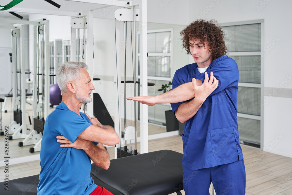 Fototapeta premium Friendly young specialist explaining how to stretching hands to his senior patient
