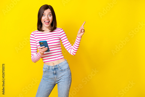 Photo of charming young girl excited sales point empty space hold gadget wear stylish striped outfit isolated on yellow color background