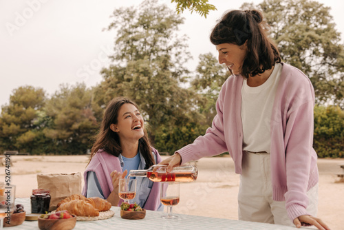 Nice young caucasian woman pours wine into glasses relaxing with her friend in nature spring. Brunettes wear casual clothes. Holiday life concept