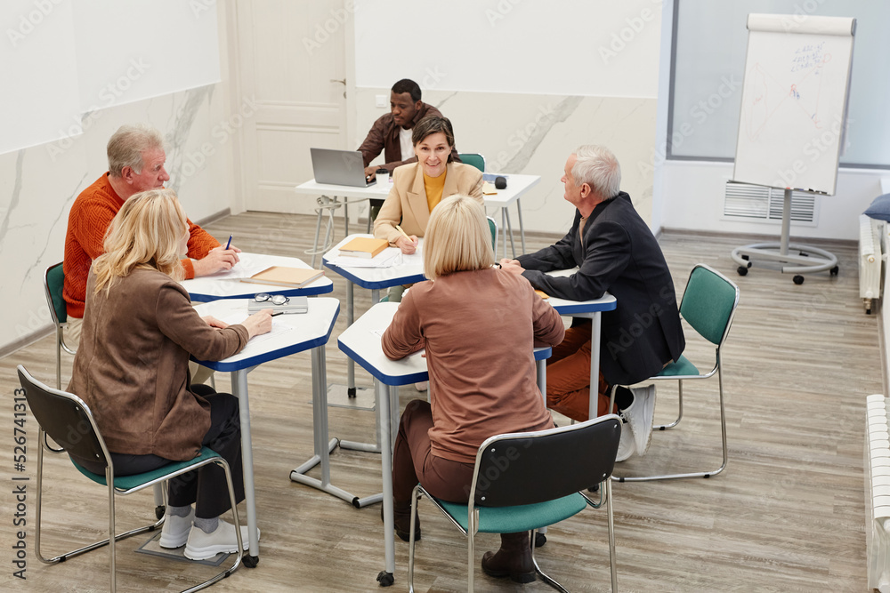Group of students sitting at desks and working in team during lesson in classroom with teacher in background