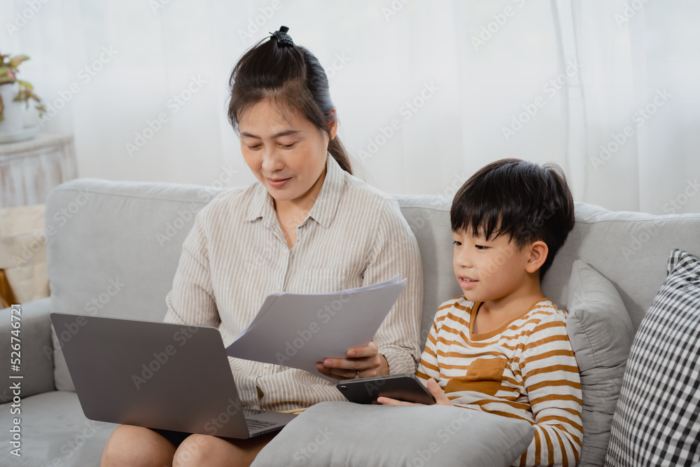 A middle-aged mother sits on the sofa intent on laptop, and her son sits on tablet next to her in the living room. The boy tried to pull the paper out of mother's hand, wanting to interest in him A middle-aged mother sits on the sofa intent on laptop, and her son sits on tablet next to her in the living room. The boy tried to pull the paper out of mother's hand, wanting to interest in him