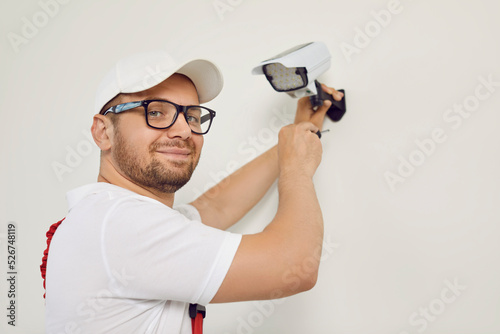 Portrait of smiling male technician worker who installing video surveillance camera. Young man in cap and glasses, who fastens surveillance camera to white wall with screwdriver, looks into camera.