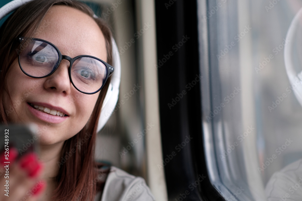 Portrait of young woman in glasses browsing mobile phone, riding on train. Close up. Pretty female using gadget while sitting in suburban train. Portrait of young woman in glasses browsing mobile phone, riding on train. Close up. Pretty female using gadget while sitting in suburban train.