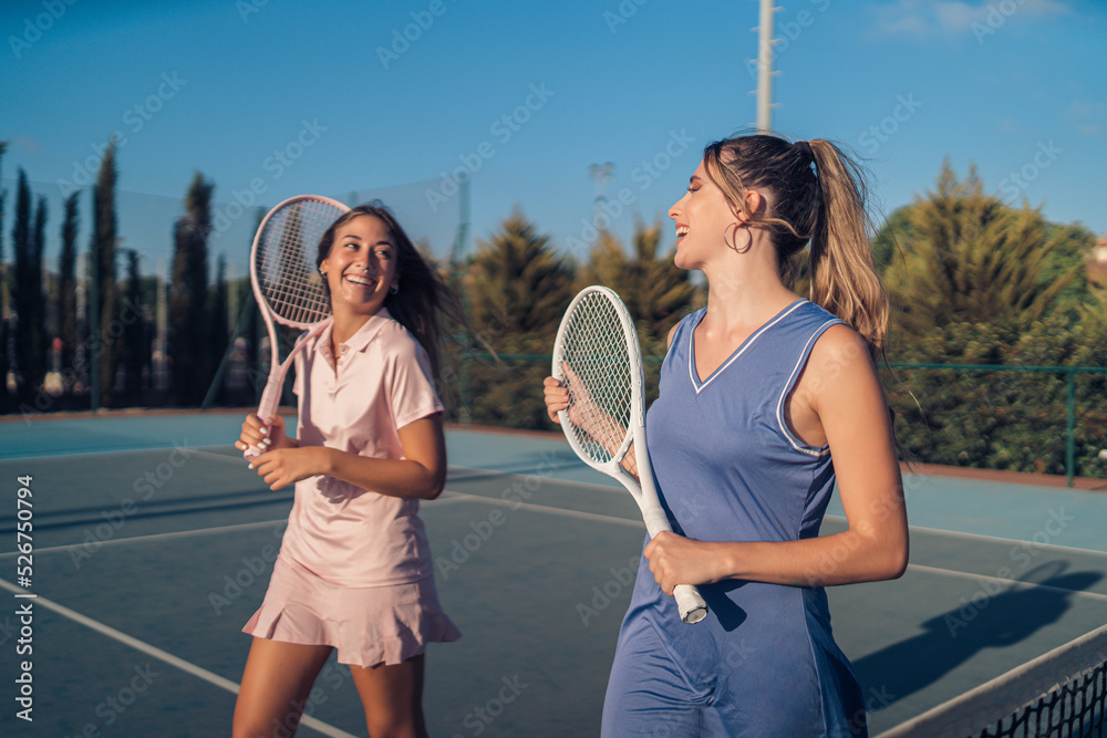 Dos chicas vestidas con ropa de tenis rosa y morada paseando juntas y haciendose selfies con smartphone en cancha de tenis
