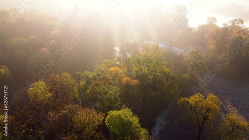 Aerial flying over trees with yellow leaves, a lake and architecture on an autumn sunny morning in a park. Bright sunlight, shining sunbeams rays and sun overexposure. Beautiful natural background.
