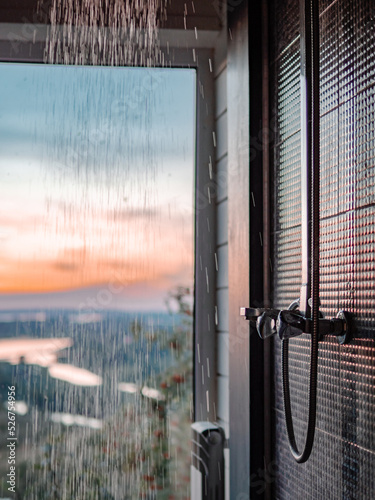 Bathing room interior with panoramic window. Splashes and drops of water from shower in bathroom with panoramic window in summer nature background at sunset.