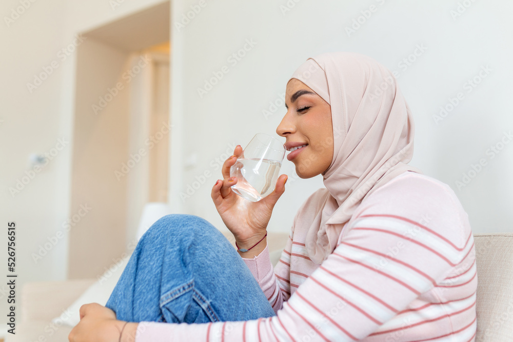 Beautiful young muslim woman with hijab drinking a fresh glass of water at home Beautiful young muslim woman with hijab drinking a fresh glass of water at home