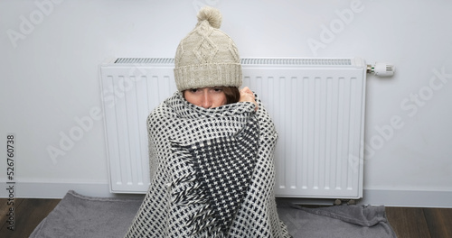 Woman cold in apartment. Young woman in sweater, scarf, hat, warm chalet sitting alone near radiator, try warming indoor. Poor heating in Europe house concept. Shaking, shivering from chilly in room 