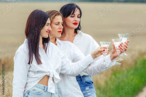 Three happy women enjoying glasses of white wine and smiling during picnic. Best friends, beautiful and stylish girls drink wine and laugh. Birthday celebration, friends toast with champagne.