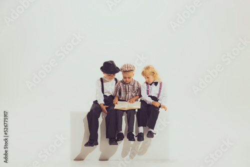 Portrait of three boys, children in stylish vintage clothes sitting together, reading book isolated over grey background photo