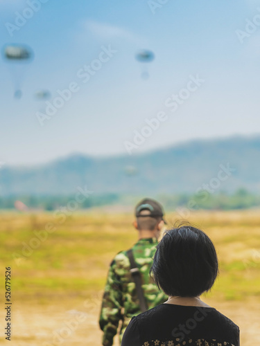 Back view of female parents look with worry and concern during parachute training from airplane for army cadet with blurred image of parachute and landscape in background. Family relationship concept. © JinnaritT