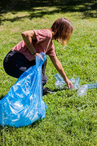Photography Young redhead woman bending down to pick up plastic bottles in public park, eco-friendly gesture