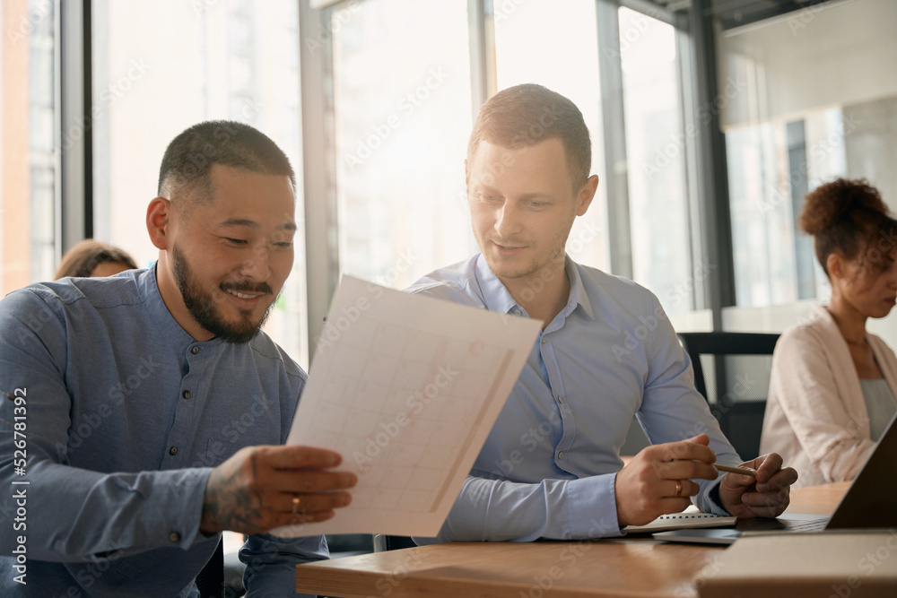 Smiling male colleague showing document to another in coworking space Smiling male colleague showing document to another in coworking space