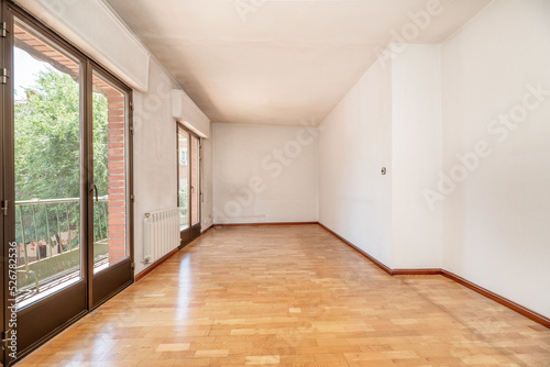 Empty living room has oak parquet floor, two balconies with bronze colored aluminum doors, plain white painted walls
