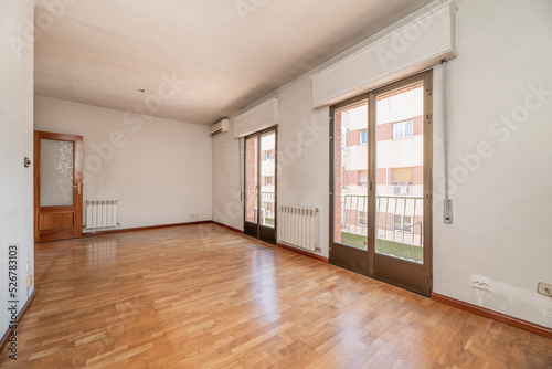 Empty living room of a residential house with oak parquet flooring, twin balconies with glass and bronze color aluminum doors with green metal railing