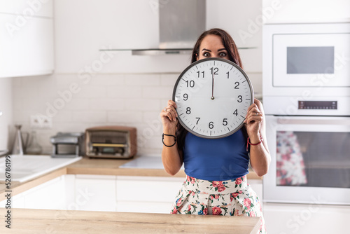 Woman stands in the kitchen holding big clock showing exactly twelve o'clock photo