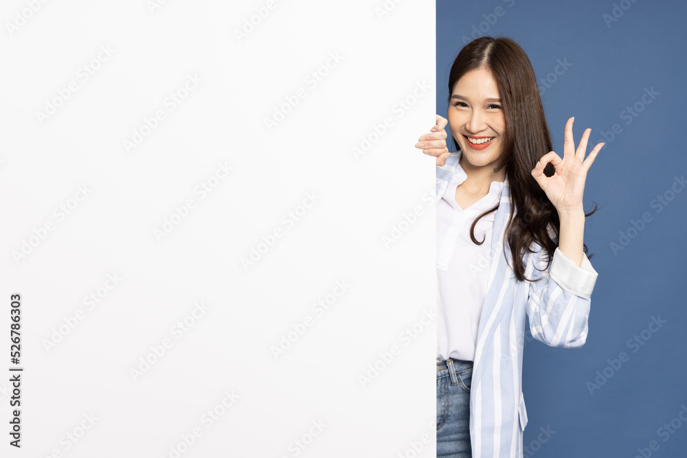 Cheerful Young Asian businesswoman is standing behind the white blank banner or empty copy space advertisement board and showing OK sign on blue background, Looking at camera Cheerful Young Asian businesswoman is standing behind the white blank banner or empty copy space advertisement board and showing OK sign on blue background, Looking at camera