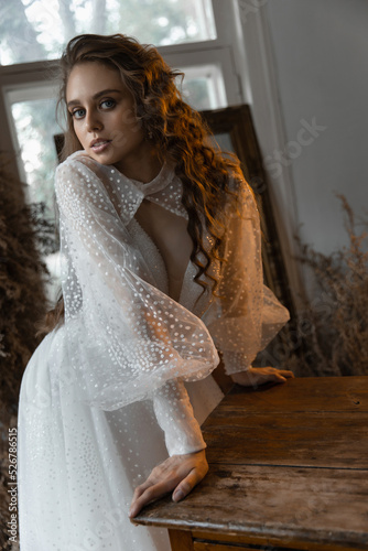 a beautiful caucasian girl bride with curly red hair stands resting against a table in a wedding dress among dry flowers and looks pathetically into the camera. fashion posing photo