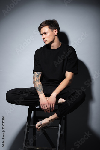 A guy of model appearance in black clothes poses on a gray studio background. Male fashion portrait