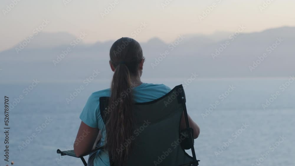 The girl is sitting on a folding chair on the edge of the mountain and looks at the sea The girl is sitting on a folding chair on the edge of the mountain and looks at the sea