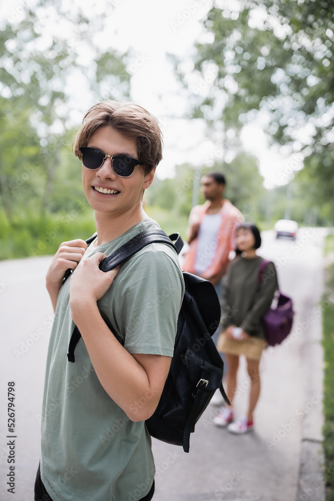 Fototapeta premium smiling traveler in sunglasses looking at camera near blurred interracial friends.
