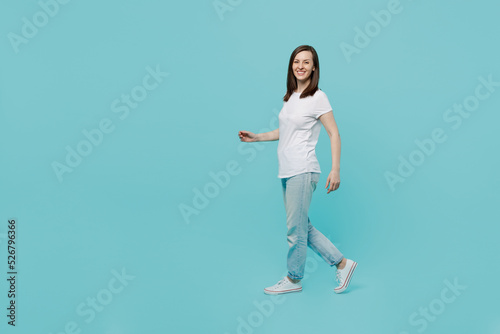 Full body side view young caucasian woman 20s she wear white t-shirt walking going strolling look camera isolated on plain pastel light blue cyan background studio portrait. People lifestyle concept.