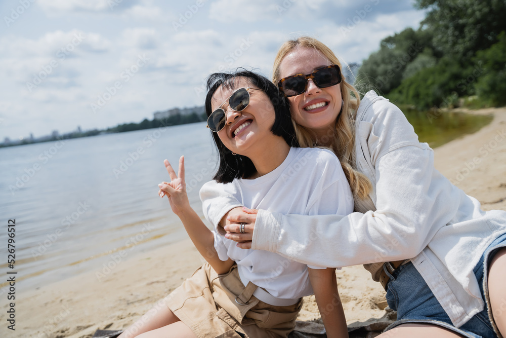 cheerful asian woman showing victory gesture near blonde friend on beach.