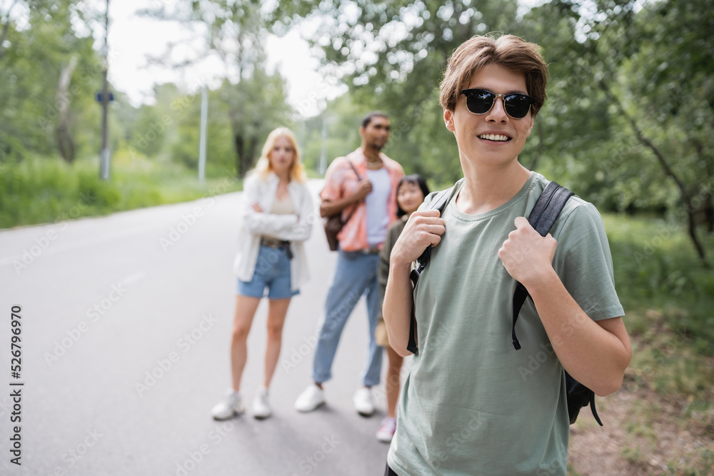 smiling man with sunglasses and backpack near multiethnic friends on blurred background. smiling man with sunglasses and backpack near multiethnic friends on blurred background.