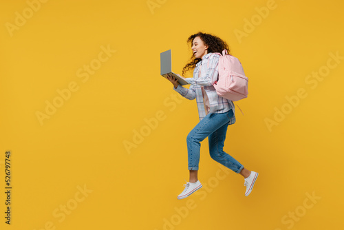 Full size side view young black teen girl student she wear casual clothes backpack bag jump high hold use laptop pc computer isolated on plain yellow background. High school university college concept