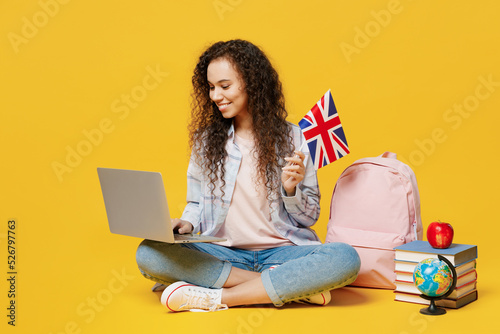 Full body young black teen girl student wear casual clothes backpack bag use work on laptop pc computer hold british flag isolated on plain yellow background. High school university college concept.