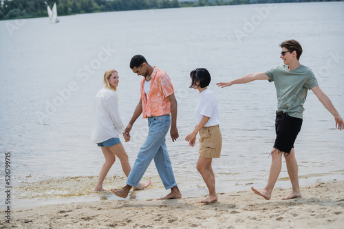 excited man walking with multiethnic friends near river and pointing with hand.