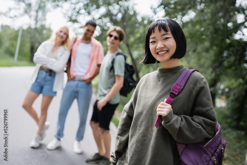 happy asian woman with backpack smiling at camera near blurred interracial friends.