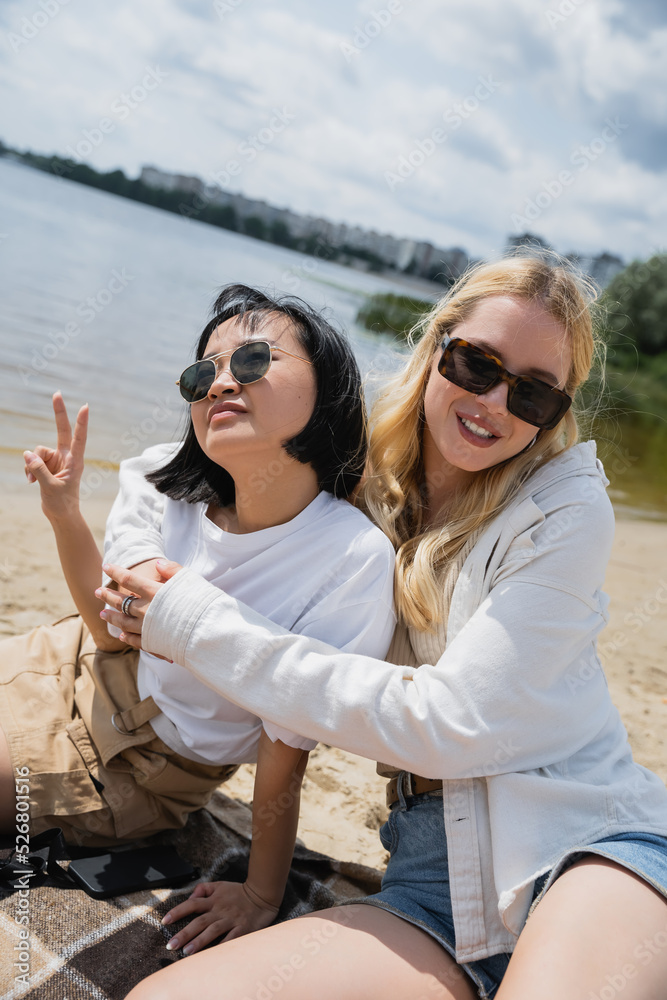 blonde woman in sunglasses embracing asian friend showing victory sign on beach.