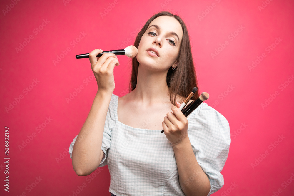 portrait of an attractive lovely young woman standing isolated over pink background, holding makeup blusher brush portrait of an attractive lovely young woman standing isolated over pink background, holding makeup blusher brush