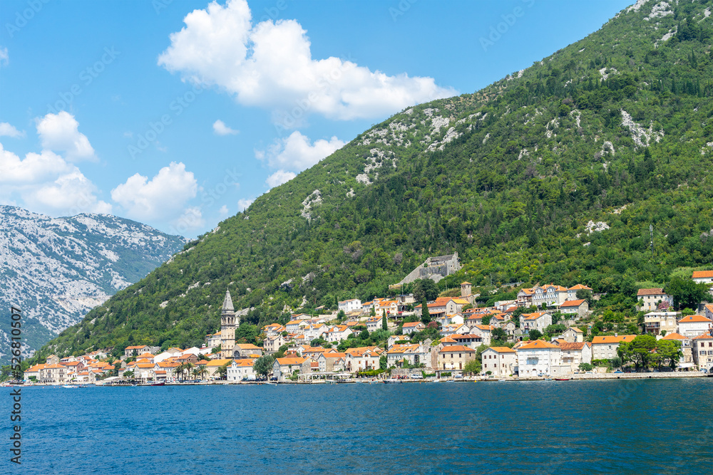 Beautiful view of seaside town at foot of mountain on sunny day on coast of sea, houses with terracotta roofs, boats and church in Montenegro, Bay of Kotor, landscape