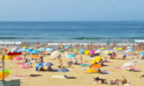 Blurred photo of ocean beach full of tourists under sun umbrellas in the summer day. Vacation background.