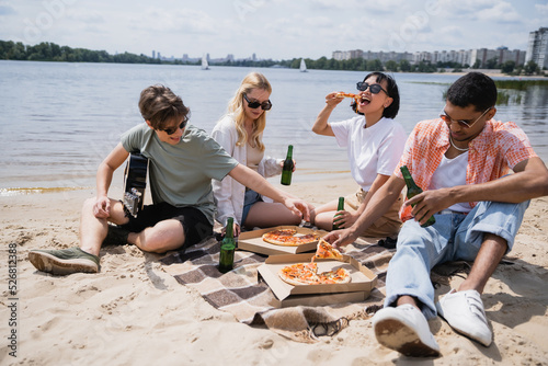 cheerful multicultural friends drinking beer and eating pizza on riverside.