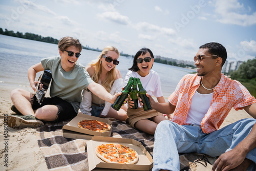 happy multiethnic friends in sunglasses toasting with beer near pizza on blanket.
