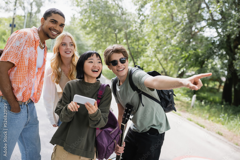 man in sunglasses pointing with finger near asian woman with smartphone and happy interracial friends. man in sunglasses pointing with finger near asian woman with smartphone and happy interracial friends.