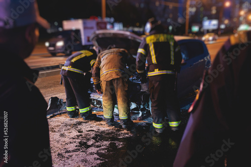 Group of fire men extinguishing and put out burning car crash after road traffic incident, fire fighting operation in the night city, firefighters with fire engine truck vehicle, emergency and rescue