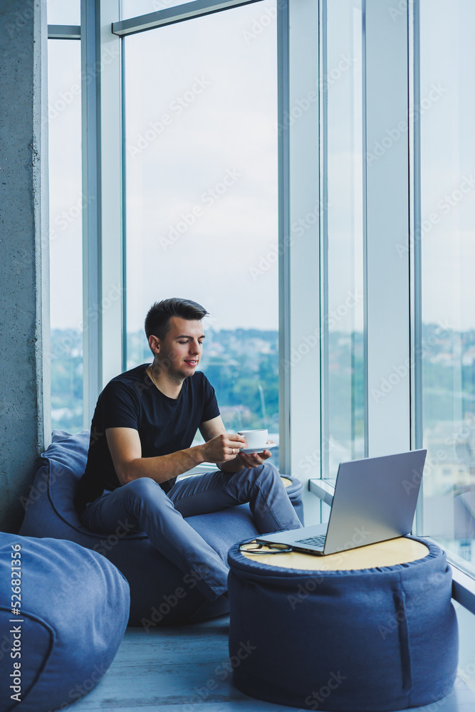 Smiling businessman looking for something and typing on a laptop and drinking fresh black coffee. The concept of a modern successful person. Young focused guy with glasses in an open office