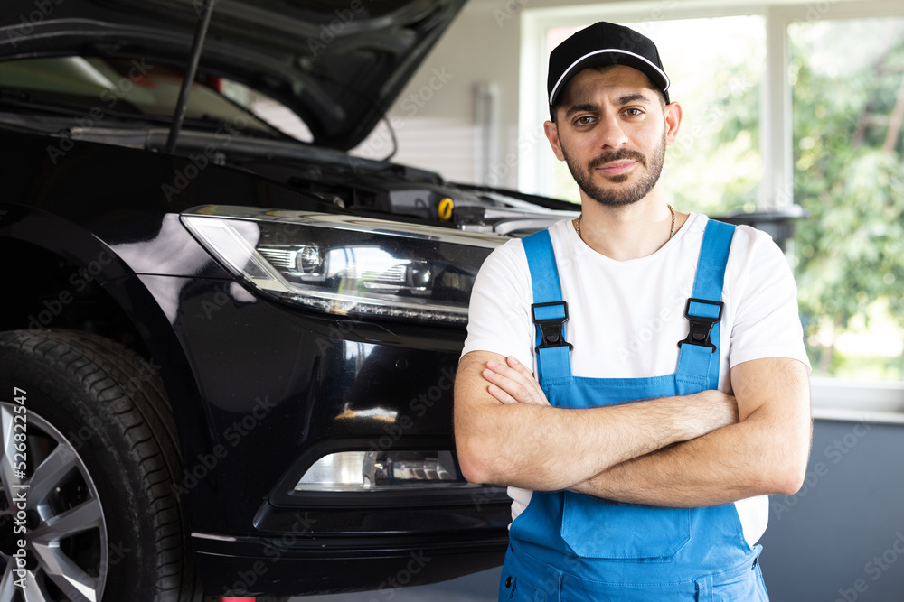 Young caucasian bearded man in blue overalls and black cap looks into camera, while smiling and crosses arms. Male car mechanic at workplace in spacious repair shop Young caucasian bearded man in blue overalls and black cap looks into camera, while smiling and crosses arms. Male car mechanic at workplace in spacious repair shop
