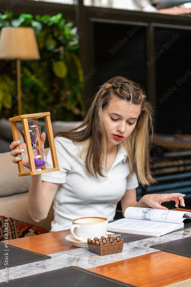 Fototapeta premium Vertical photo of young lady holding hourglass and reading her book
