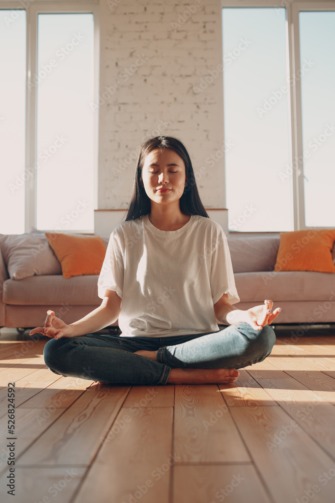 Asian woman doing yoga and zen like meditation lotus pose in casual wear at indoor living room apartment with natural sun light.