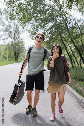 excited interracial couple with backpacks and guitar laughing while walking on road. © LIGHTFIELD STUDIOS