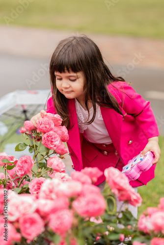 charming little girl in a bright pink suit sniffs a rose bush. card, banner