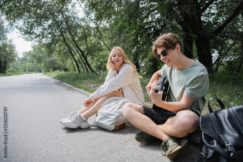 man sitting on road with crossed legs and playing guitar to blonde girlfriend.