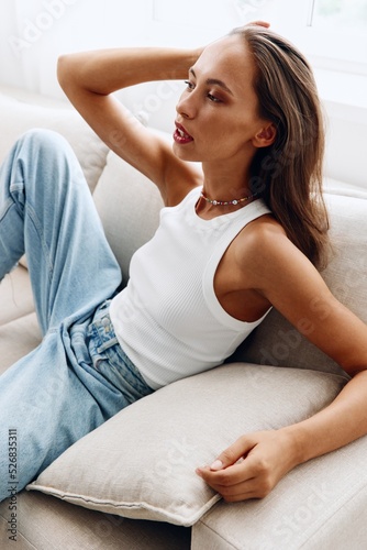 Beautiful young woman lying on the couch at home in a relaxed pose, fixing her long hair in a white T-shirt and blue jeans