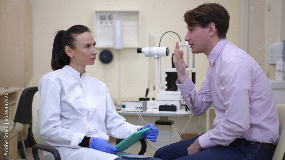 Side view portrait of Caucasian man talking to oculist complaining bad eyesight as concentrated professional woman listening sitting with digital tablet indoors. Patient and doctor on appointment Side view portrait of Caucasian man talking to oculist complaining bad eyesight as concentrated professional woman listening sitting with digital tablet indoors. Patient and doctor on appointment